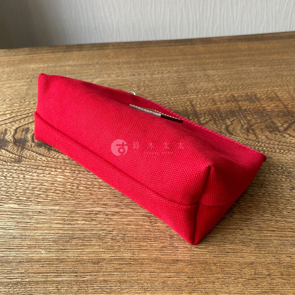 紅色石洗帆布製成的小型收納包，包體呈現長方形，一側有拉鍊，質感粗獷，置於木紋桌面。