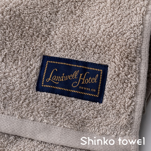 米色織紋毛巾，帶有藍色標籤，上面印有金色字體的「Landwell Hotel TOWEL CO.」字樣。
