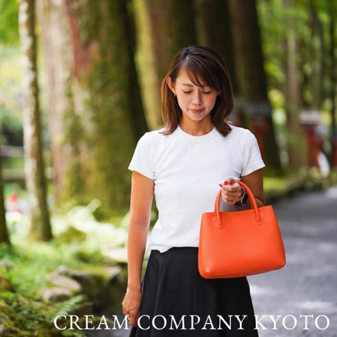 一位女性手持亮橙色手提托特包，包身為皮革材質，設計簡約。背景為自然景觀，有樹木與小徑。