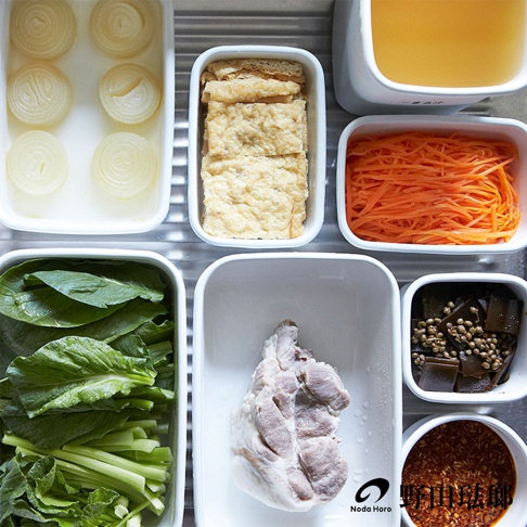 多個白色長方型淺型琺瑯保存容器，其中裝有切片的洋蔥、炸豆腐、切絲的紅蘿蔔、綠葉蔬菜、豬肉塊，以及醬料和香料。