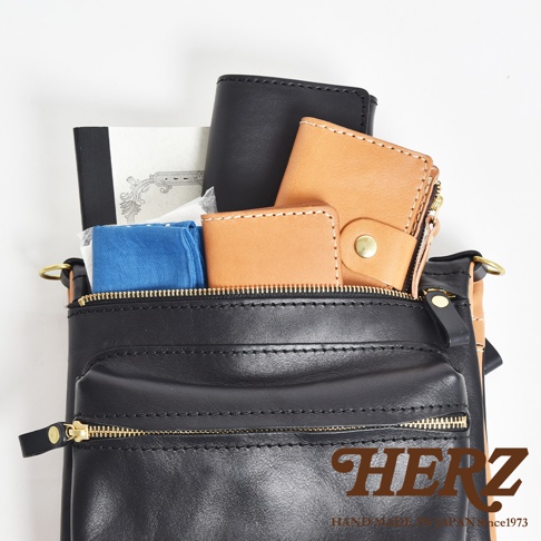 黑色皮革翻轉直式斜背包，內有棕色皮夾、筆記本和藍色布料，搭配金色拉鍊和扣件。