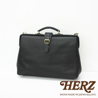 黑色皮革軟式杜勒斯包，包身呈現方形，頂部有提把，正面設有皮質束帶和黃銅金屬扣環，風格簡約。