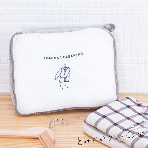 白色網狀方型洗衣袋，印有黑色「TOMIOKA CLEANING」字樣及一件西裝圖案，側面有灰色網格及拉鍊，下方疊放著一件格子襯衫。
