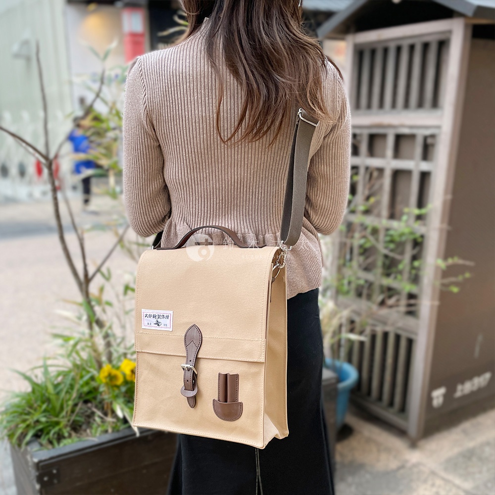 米色純棉帆布製成的直式側背包，配有棕色皮革翻蓋、扣環和提把，可斜挎或手提。