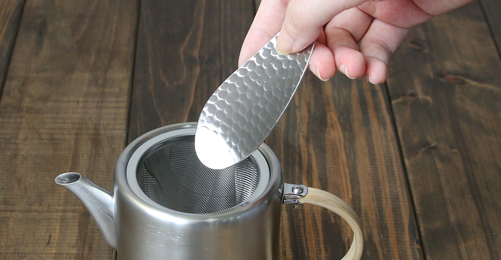 手持一個銀色不鏽鋼製的茶勺，茶勺表面有錘紋紋理，勺頭呈葉片狀，正準備放入一個帶有細網濾茶器的銀色金屬茶壺中。