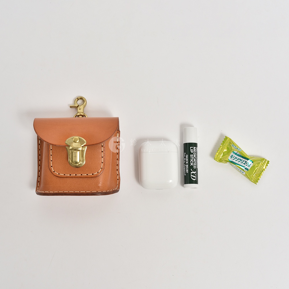 職人手作皮革耳機收納包，淺棕色植鞣皮革，附黃銅金屬釦和掛鉤，旁邊擺放白色無線耳機、藥用潤唇膏和綠色糖果。