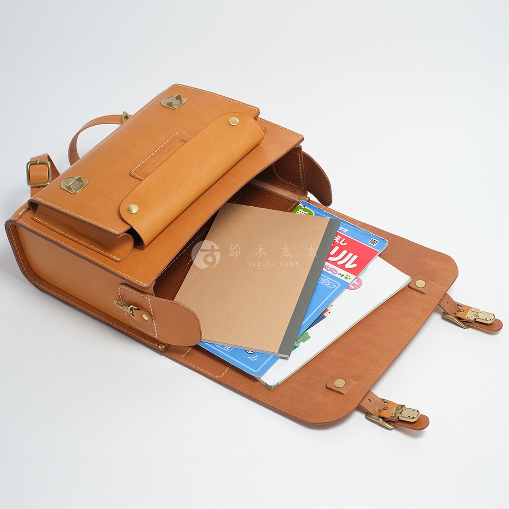 職人手作皮革橫式書包，打開的包蓋內可見兩本筆記本，其中一本為棕色封面的筆記本，另一本為藍色封面的筆記本，筆記本上印有日文文字。
