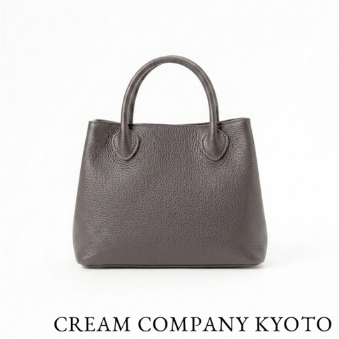 一個由日本京都品牌 CREAM COMPANY KYOTO 出品的深灰色皮革手提托特包，採用紋理細緻的牛皮革製作，包身造型簡約優雅，正面呈現飽滿的皮革質感與精緻的提把縫線細節，背景為純白色。