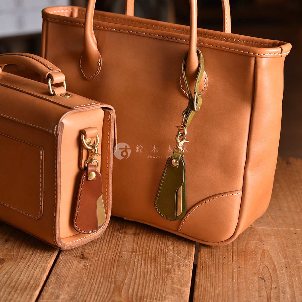 職人手作皮革隨身金屬鞋拔，以鞣製過的牛皮製成，呈現自然棕色調。鞋拔結合了皮革與金屬材質，部分款式帶有綠色皮革裝飾，並配有金屬掛環，可掛於包袋上。