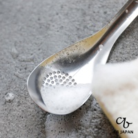 CB JAPAN 日本製不鏽鋼瀝汁匙，水珠沾附在金屬勺面上，勺內有泡沫，背景是灰色紋理桌面。