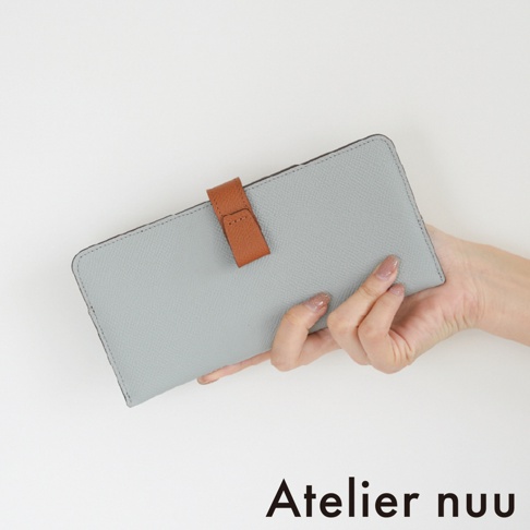 一隻手持著 Atelier nuu 出品的 noble 皮革雙折長夾，長夾主體為簡約的淺灰色細紋皮革，中央配有顯眼的棕色皮革扣環，整體呈現細緻且優雅的設計風格。
