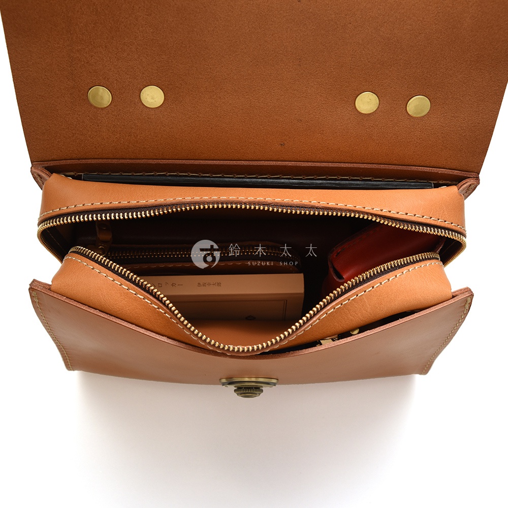 日本HERZ職人手作皮革多功能收納單肩包，內部拉鍊打開，展示出皮革內裡、一本較厚的書本、一個小巧的紅色皮夾，以及印有SUZUKI SHOP字樣的標籤。