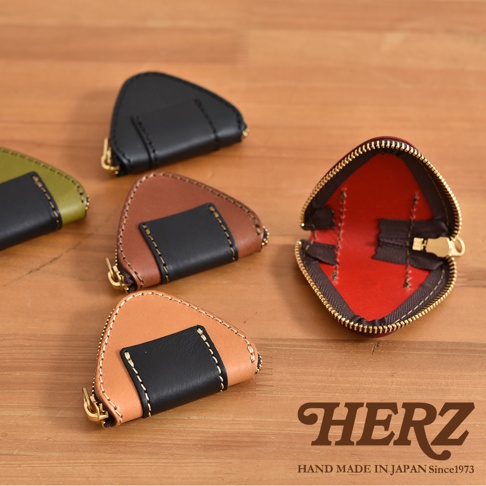 木質桌面上放置了五個日本HERZ職人手作的三角飯糰造型皮革零錢包，包含黑色、棕色、淺棕色及綠色拼接款式，右側展示一個打開的紅色內裡零錢包，呈現出精緻的皮革縫製細節與拉鍊設計。