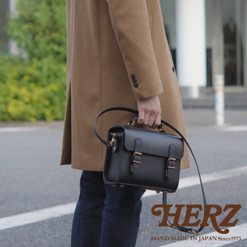 黑色皮革迷你兩用包，配有頂部提把和可拆卸肩帶，採用雙搭扣閉合設計，呈現職人手作的極簡風格。