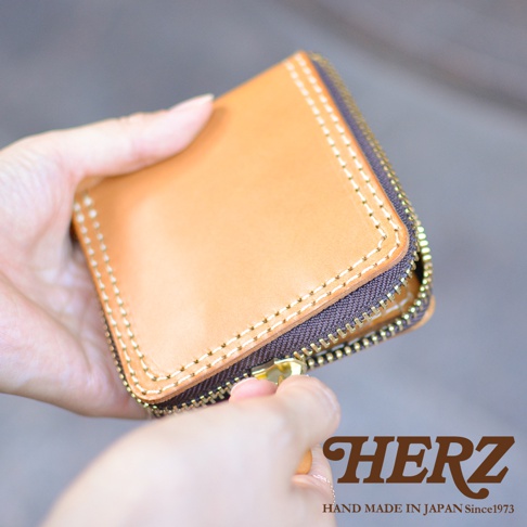 這是一雙手正拿著一個日本HERZ職人手作的淺棕色皮革短夾，皮夾四周飾有粗縫線，並搭配深棕色拉鍊設計，右下角印有HERZ品牌字樣與產地標示。