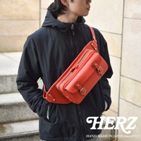 職人手作皮革騎士隨身斜背包，採用鮮豔的紅色皮革製成，包款具有雙層拉鍊主袋、前方翻蓋口袋，並以銅釦和皮革繫帶固定，斜背於男性模特兒身上。