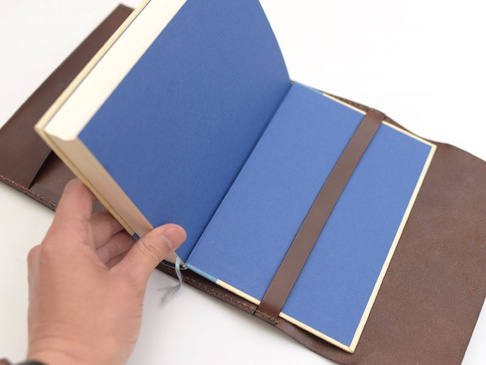 深棕色皮革翻蓋記事本書套，內襯藍色紙張，附有皮革固定帶，左手正在翻開書本。