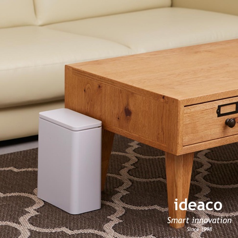 白色簡約方形垃圾桶，設有按壓式開啟的白色頂蓋，置於木質桌子旁的地毯上。