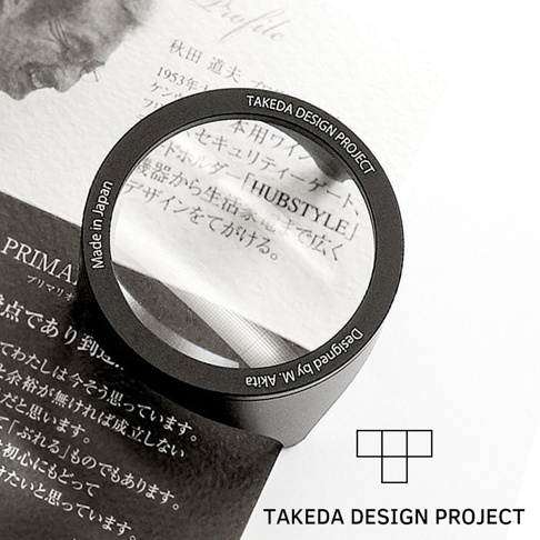 黑色鋁製閱讀放大鏡，鏡框上刻有「Made in Japan」及「TAKEDA DESIGN PROJECT」字樣，鏡片放大文字，整體呈現簡約設計。