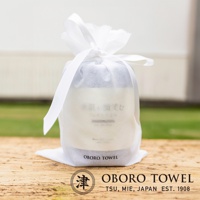 白色半透明網狀束口袋包裝著一條淺灰色毛巾，上面印有日文「潤澤素肌微笑速吸毛巾」及英文「OBORO TOWEL」。束口袋以白色緞帶繫緊，整體呈圓柱狀，放置於木質桌面上，背景為模糊的綠色草地與白色牆壁。
