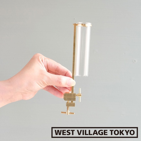 一只手持著黃銅可夾式玻璃花器，花器由黃銅支架和透明直筒形玻璃瓶組成，玻璃瓶帶有垂直紋理。