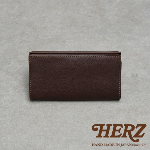 這是一個由日本品牌HERZ推出的職人手作皮革長夾，呈現深棕色的皮革質感，外觀採用簡約的長方形摺疊設計，邊緣有精細的車縫線裝飾，右下角印有棕色的HERZ品牌標誌及其創立年份。