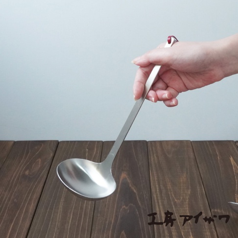 一只手正握著一個不鏽鋼湯勺，湯勺勺頭為圓弧形，勺柄細長，整體為銀白色金屬光澤。