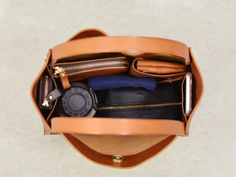 職人手作皮革Organ方形托特包，淺棕色皮革，內部展示收納物品，包含一個黑色水瓶、一個深藍色織物、一個黑色拉鍊小包、一個棕色長夾、一個卡夾和一部白色手機。