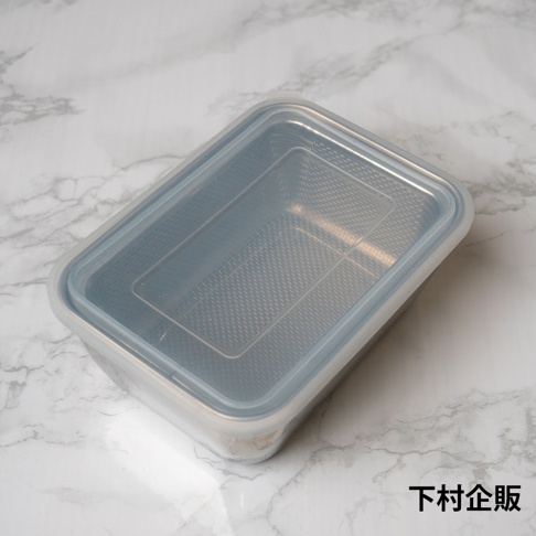 一個長方形的下村企販不鏽鋼多功能調理保存盒放置在大理石紋桌面上，容器由不鏽鋼製成，頂部配有透明塑膠蓋，整體呈現簡約實用的廚房收納風格，視角由上往下俯拍。