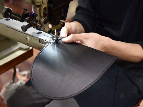 職人正在使用工業縫紉機縫製一個黑色的皮革翻蓋迷你單肩包，展示精緻的車縫細節。