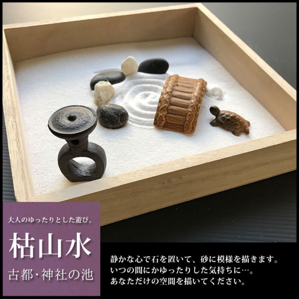 日式枯山水模型，木質淺盤盛裝白色砂礫，砂礫上佈有耙痕紋路。盤中置有深色仿古燈飾、數顆黑白石子、一座深棕色木紋橋樑模型，以及一隻淺棕色小烏龜模型。