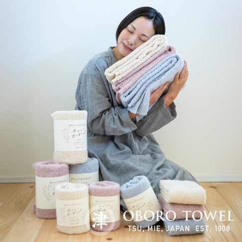 一位女士抱著一疊有機棉浴巾，顏色有米白、淡粉和淺藍。旁邊擺放著幾捲不同顏色的浴巾，皆為有機棉材質，包裝上印有日文和品牌標誌。