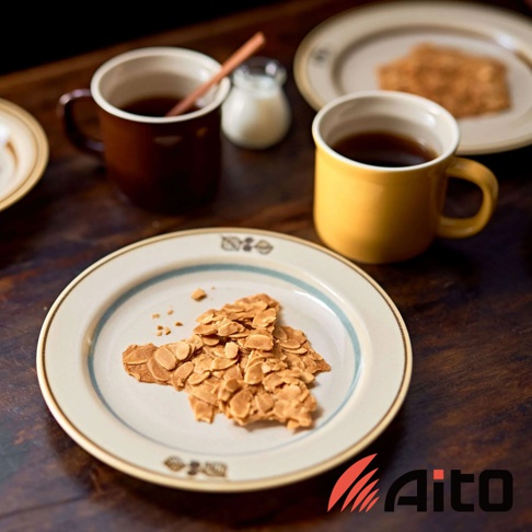 淺色美濃燒盤，盤緣有藍色條紋及葉紋圖案，盤中擺放著一片杏仁脆餅。背景有兩只馬克杯，一只深棕色，一只鵝黃色，皆裝有黑色飲品。