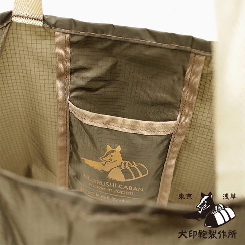 犬印鞄環保折疊收納購物袋，採用綠棕色格紋尼龍材質，內側有同色系尼龍小口袋，上面印有金色的狐狸及行李圖案，並標示「INUJIRUSHI KABAN made in Japan Pocket Tote」。