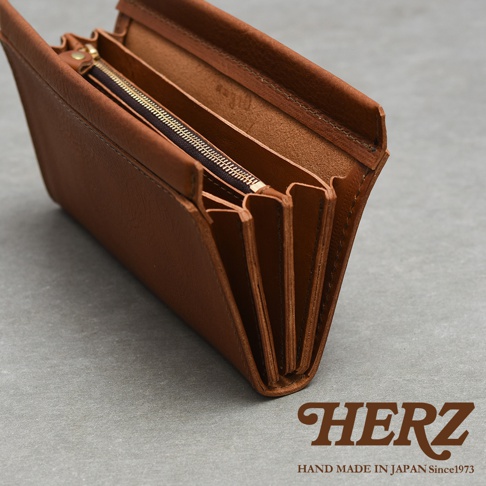 一個淺棕色的日本HERZ職人手作皮革長夾，呈現半開視角，展示出內部多層次的風琴式風琴摺夾層以及帶有黃銅拉鍊的零錢袋，整體設計簡約厚實，右下角印有HERZ品牌商標與日本手工製作的文字標示。