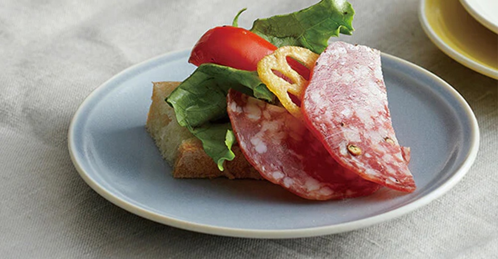 淺藍色美濃燒配菜盤上，擺放著一片厚實的麵包，搭配生菜、小番茄、炸蓮藕片和一片切片香腸。