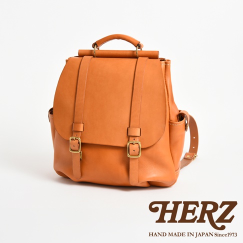 一款日本HERZ手工製作的棕色皮革後背包，呈現經典屋瓦風設計，正面配有兩條皮帶扣環，包身上方設有堅固的皮革提把與金屬結構，整體簡約耐用，下方印有HERZ品牌標誌與Hand Made In Japan字樣。