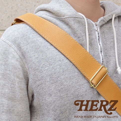 身穿灰色連帽衫的人，肩上背著一個裝有棕色皮革肩帶的包，上面印有HERZ和HAND MADE IN JAPAN Since1973的字樣。