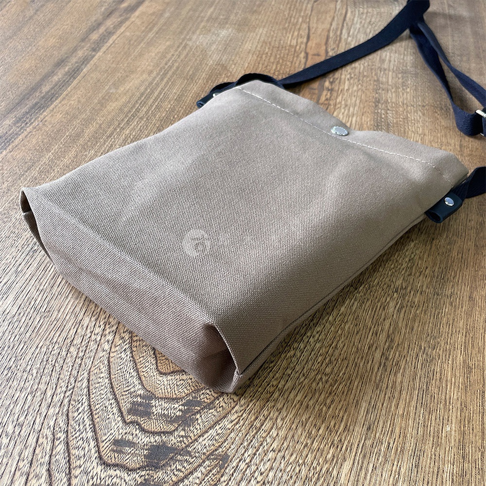 一個米褐色的帆布平板收納側背包，斜背帶為黑色，包包上有一個銀色按扣，放置於木紋桌面上。