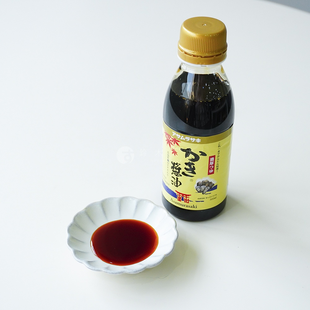 白色桌面上放置著一瓶裝在透明塑料瓶中的朝紫廣島牡蠣醬油，瓶蓋為金色，瓶身貼有標示品牌名與牡蠣圖案的黃色標籤，左側擺放一個盛有少量深褐色醬油的白色波浪狀淺碟。