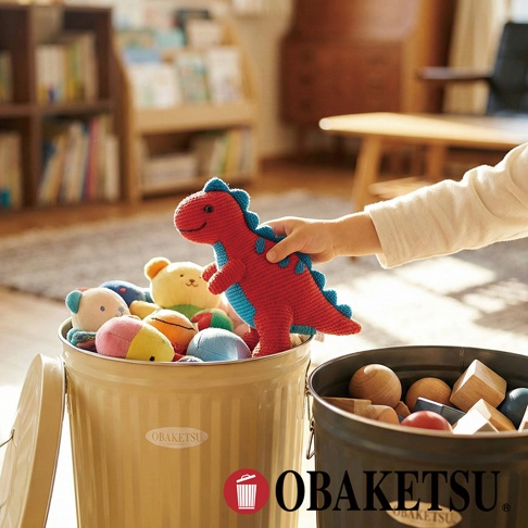 一個裝滿可愛玩具的黃色 OBAKETSU 垃圾桶，一個小朋友的手正從桶裡拿起一個紅藍色的編織恐龍玩偶。
