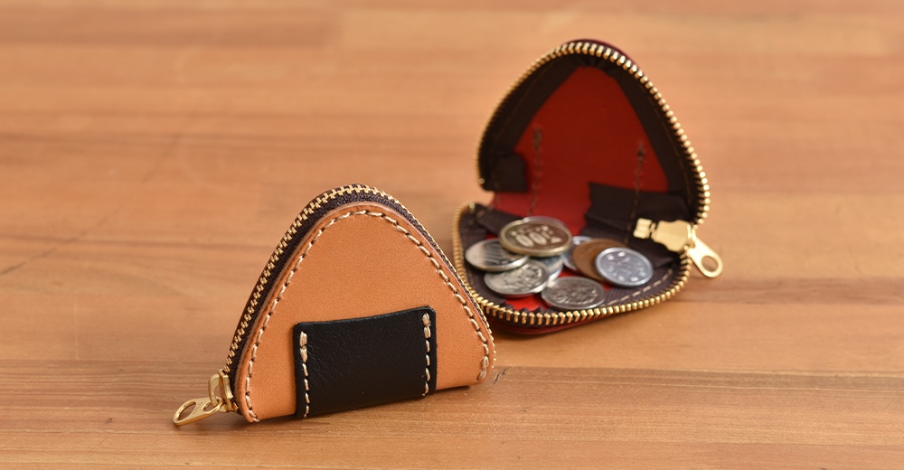 兩個日本職人手工製作的皮革三角飯糰造型零錢包放置在木質桌面上，前方一個閉合的淺棕色皮革零錢包帶有黑色皮革裝飾，後方另一個打開的零錢包內部為紅色，裝有數枚硬幣。