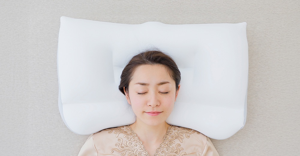 一名女子閉著眼睛仰躺在淺色床面上，頭部舒適地枕在一個白色的人體工學枕頭上，從正上方俯視的角度呈現。
