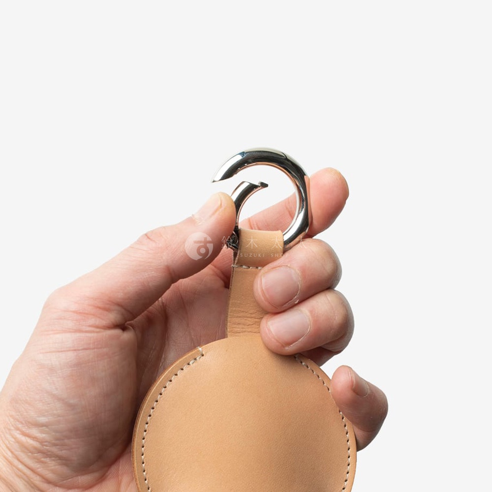 左手握著一個光滑的銀色鉤環，下方連接著一個米色皮革製成的圓形收納包，包邊緣有縫線細節。