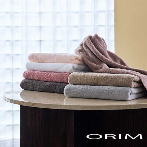桌面上整齊堆疊著多條 ORIM 日本製頂級今治認證毛巾，包含淺粉、白色、乾燥玫瑰色、深灰、駝色與淺灰色等多種質感色調，柔軟的毛巾層疊出厚實澎潤的視覺效果，右上方垂墜的毛巾展示了細膩的棉質紋理，下方標註品牌名稱 ORIM。