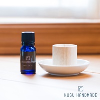 藍色小玻璃瓶裝有樟樹精油，瓶身貼有黑色標籤，寫有「KUSU HANDMADE CAMPHOR OIL」。旁為一個淺白色圓形陶瓷盤，盤上放置一個淺木色、開口朝上的木質擴香筒。