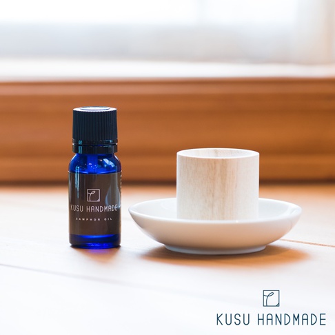 藍色小玻璃瓶裝有樟樹精油，瓶身貼有黑色標籤，寫有「KUSU HANDMADE CAMPHOR OIL」。旁為一個淺白色圓形陶瓷盤，盤上放置一個淺木色、開口朝上的木質擴香筒。