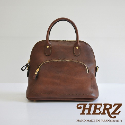 職人手作經典貝殼造型皮革手提包，採用深棕色皮革，配有金色拉鍊與金屬铆釘，包身設有拉鍊前袋，設計簡約實用。