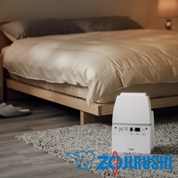 白色多功能智慧烘乾機置於臥室地板的毛毯上，旁邊是木製床架和米白色床單，床頭有兩個灰色枕頭。