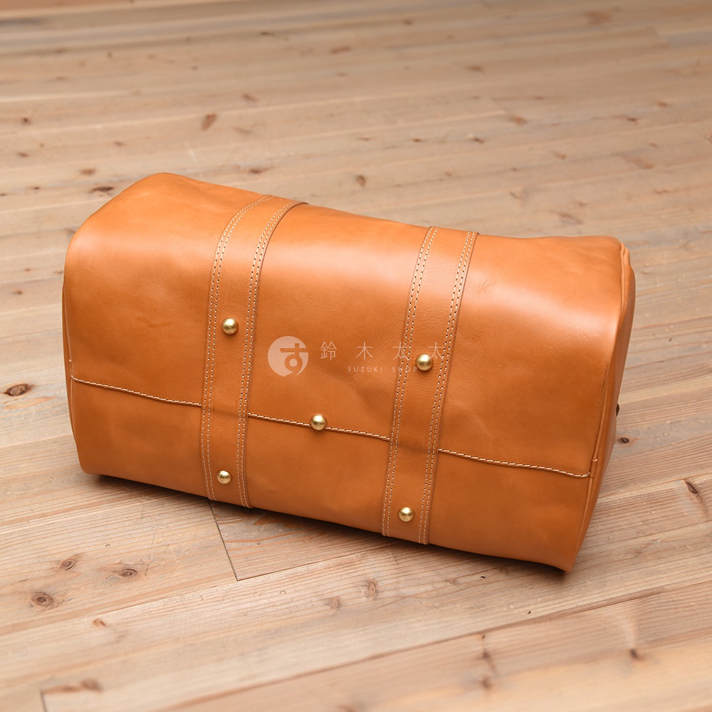 淺棕色皮革製成的圓筒形波士頓包，帶有提把和金屬鉚釘裝飾，置於木質地板上。